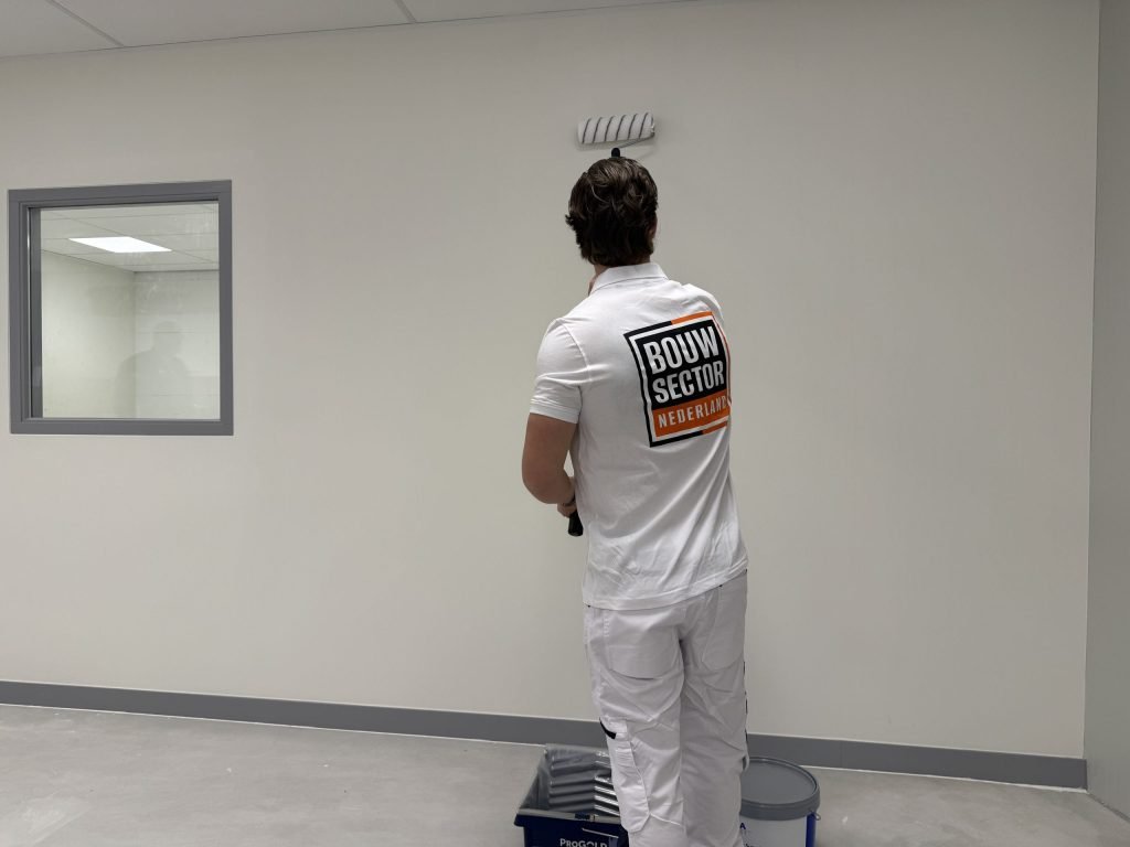 Schilder in wit werktenue saust een lichte wand, met verfbak en emmer aan zijn voeten