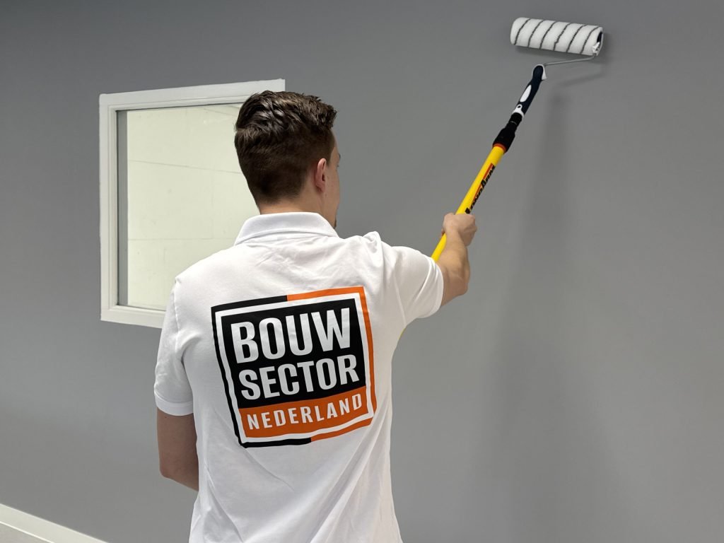 Schilder met Bouwsector Nederland-logo op de rug zet een muur in een grijze kleur met de roller, naast een binnenraam