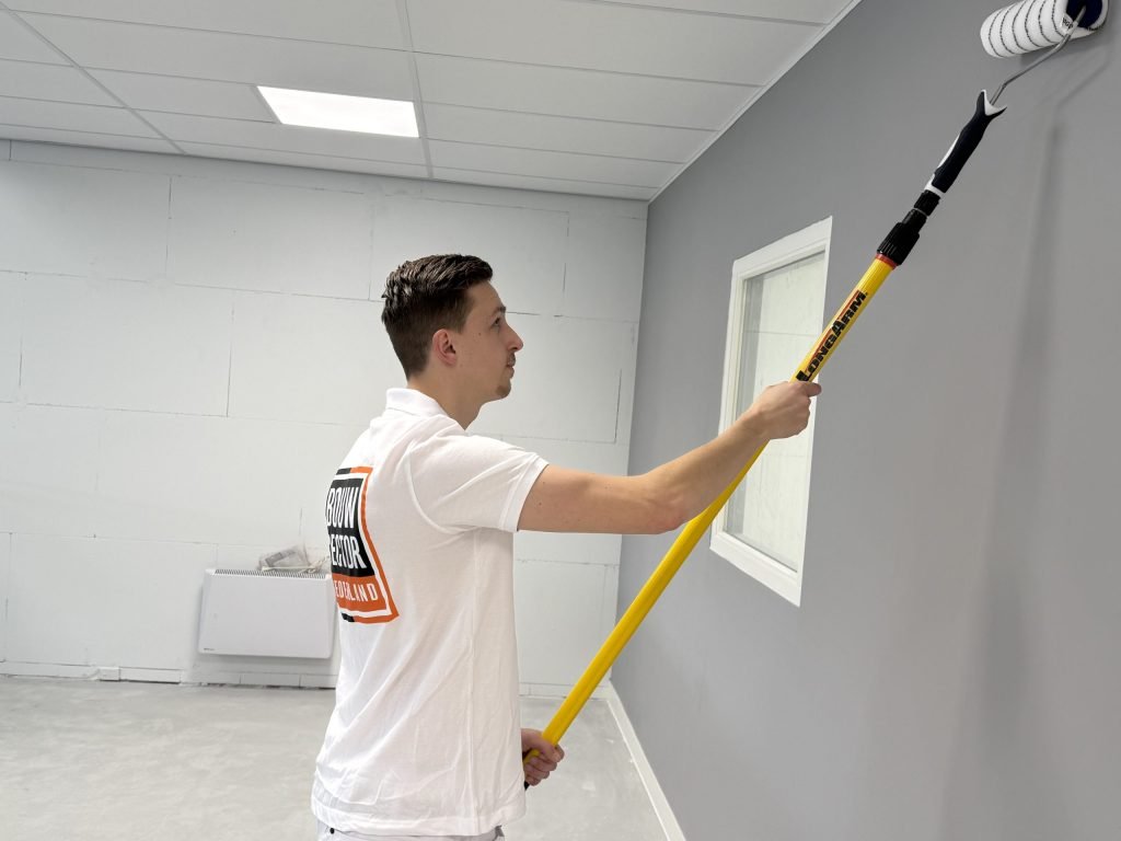 Schilder in wit poloshirt rolt met een lange verlengsteel een grijze wand bij een raam, met een systeemplafond erboven