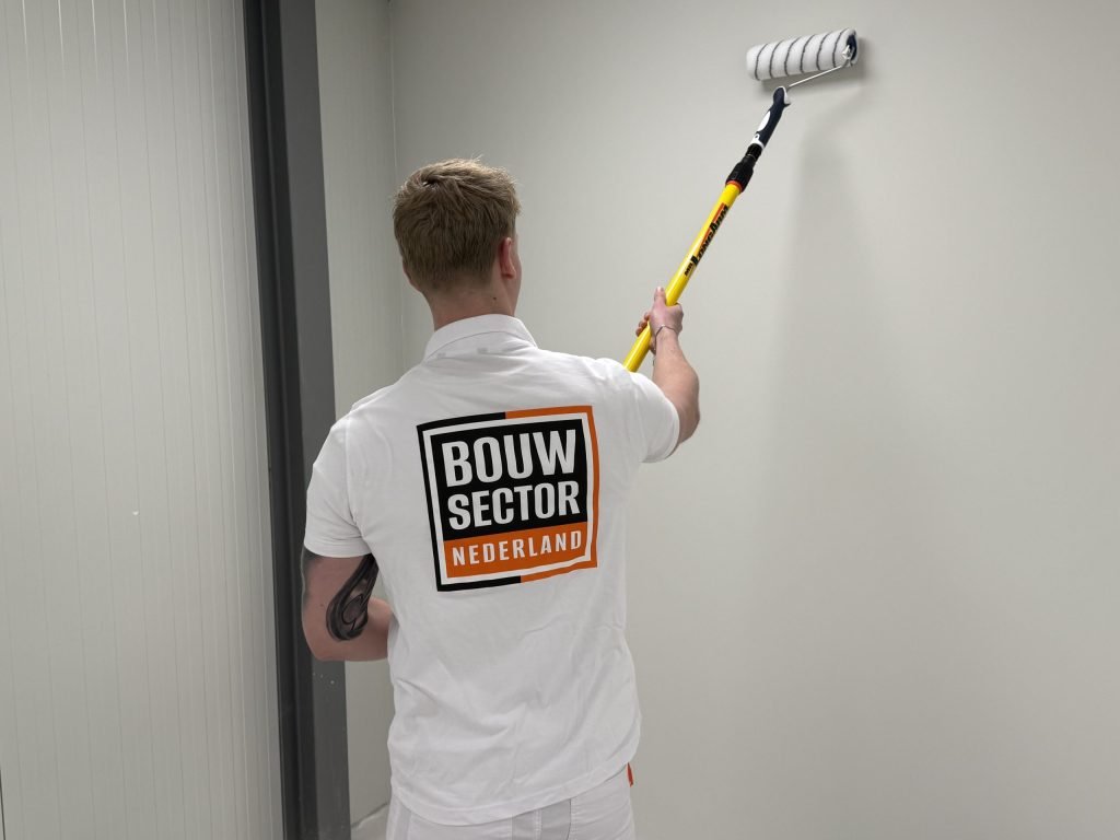 Schilder met het Bouwsector Nederland-logo op de rug rolt een lichte wand met een roller aan een gele steel