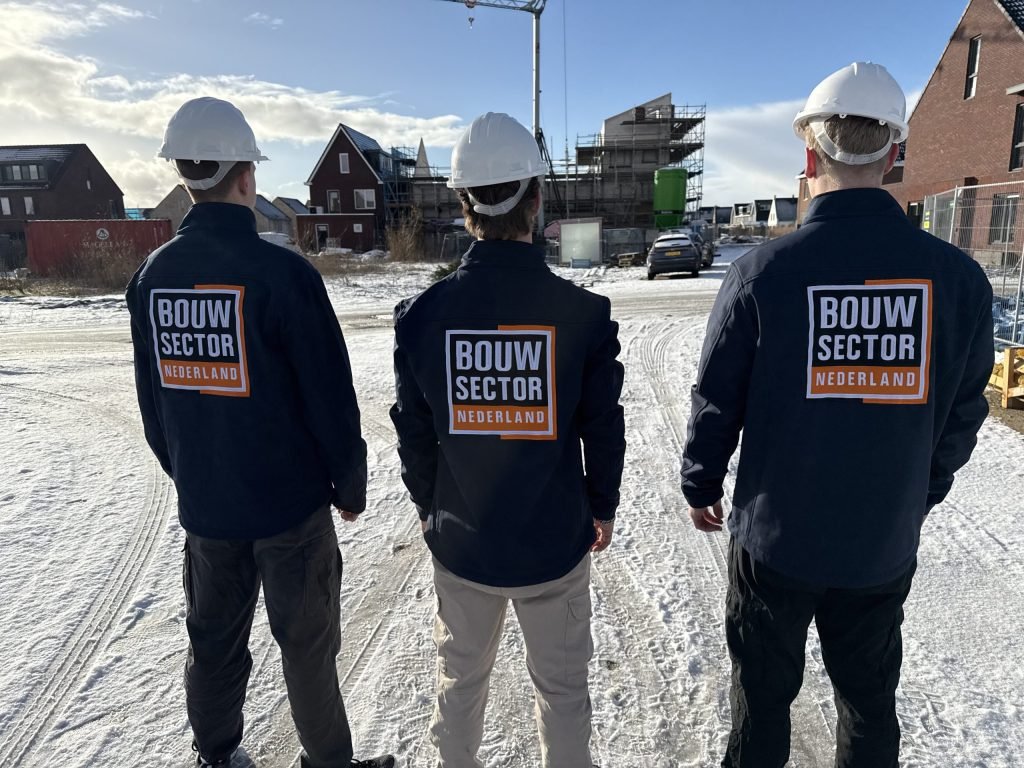 Drie vakmensen van Bouwsector Nederland met witte helm op een besneeuwde bouwweg tussen nieuwbouwwoningen