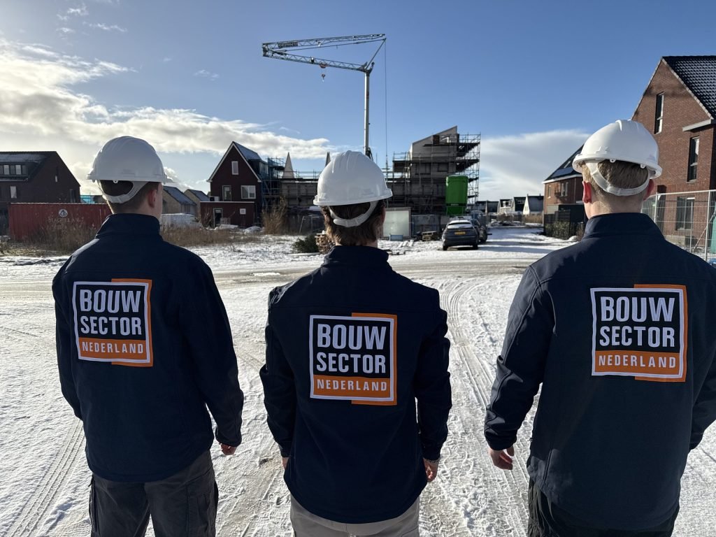 Drie vakmensen van Bouwsector Nederland met witte helm staan op een besneeuwde weg tussen huizen in aanbouw